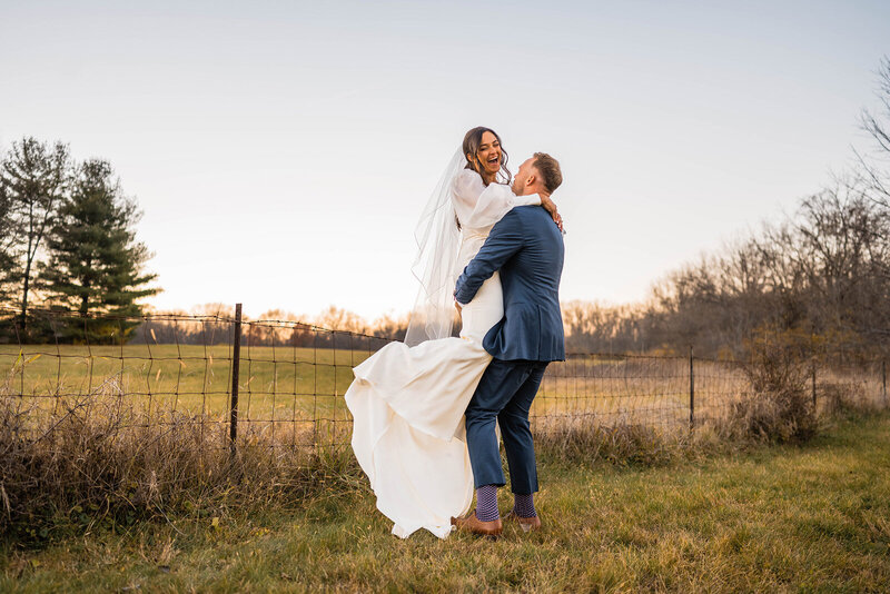 Groom picks up a laughing bride while spinning with her in a field at sunset.