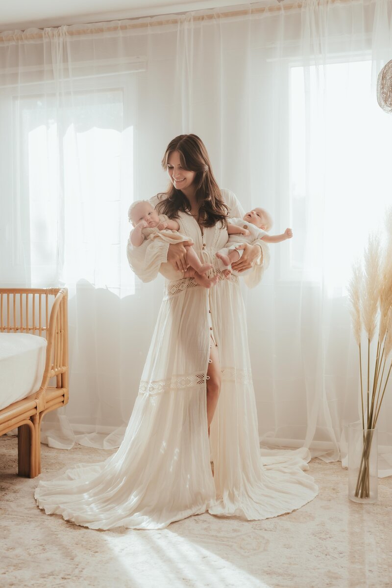 A mom in a beautiful white dress holding her twin babies in her arms.