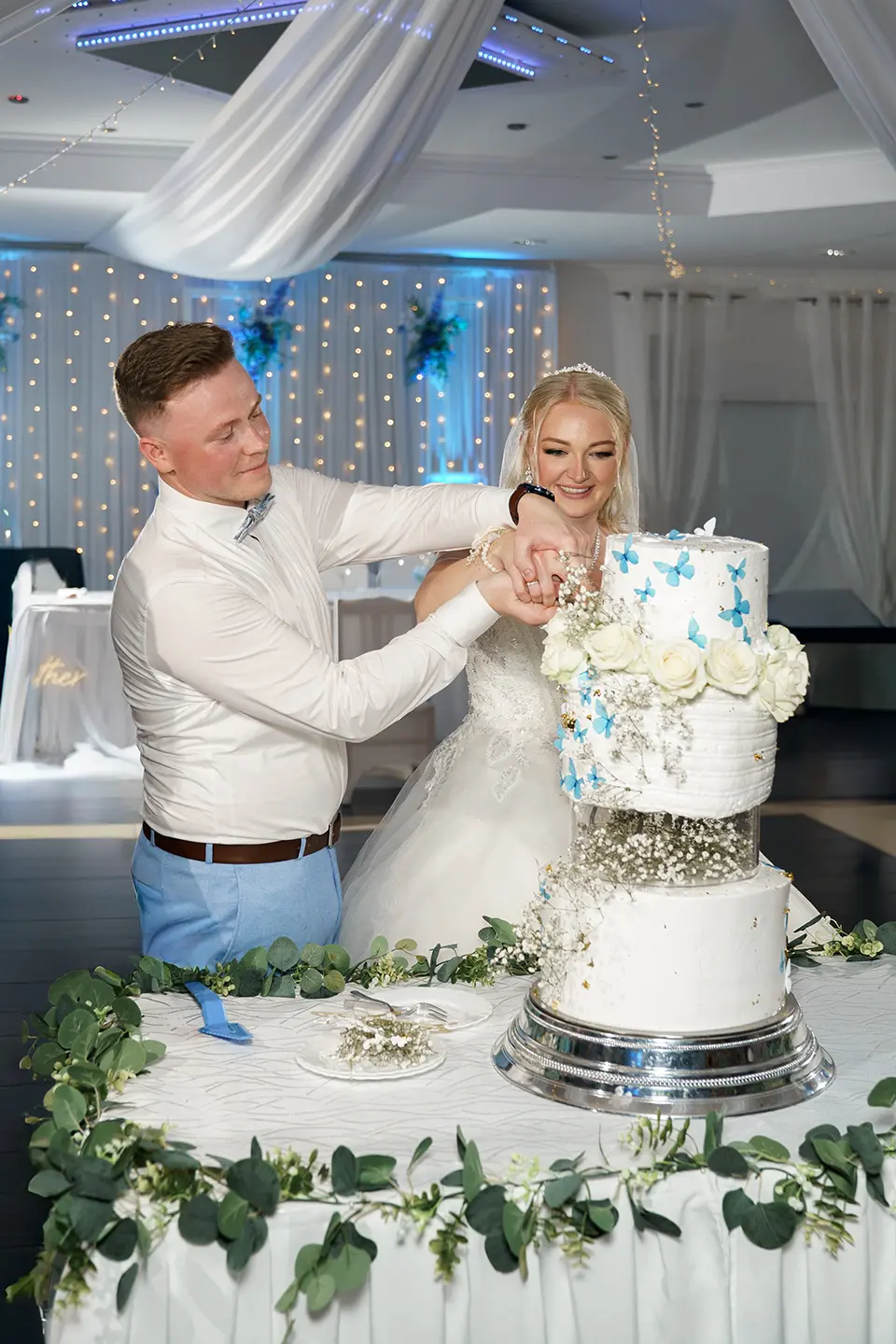Braut und Bräutigam schneiden gemeinsam eine weiße Torte mit blauen Schmetterlingen; Lichter und Eukalyptus schmücken den Tisch – reportagefotografie hochzeit und fotograf für hochzeitsfeier