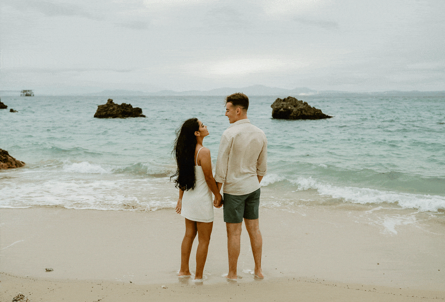 J & B holding hands facing the ocean kissing