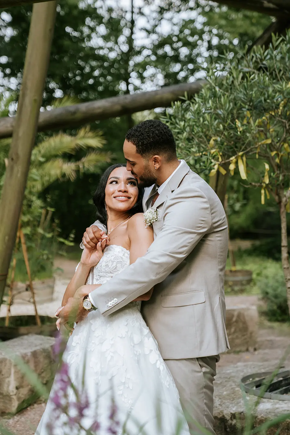 Der Bräutigam hält die Braut liebevoll im Arm, Holzstreben und Grün schaffen einen intimen Rahmen. Hochzeitsfotografie mit Stil und reportagefotografie hochzeit harmonisch vereint