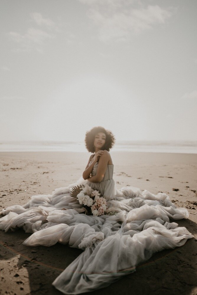 Bride sitting on the beach with a flowing gown and bouquet under soft sunlight