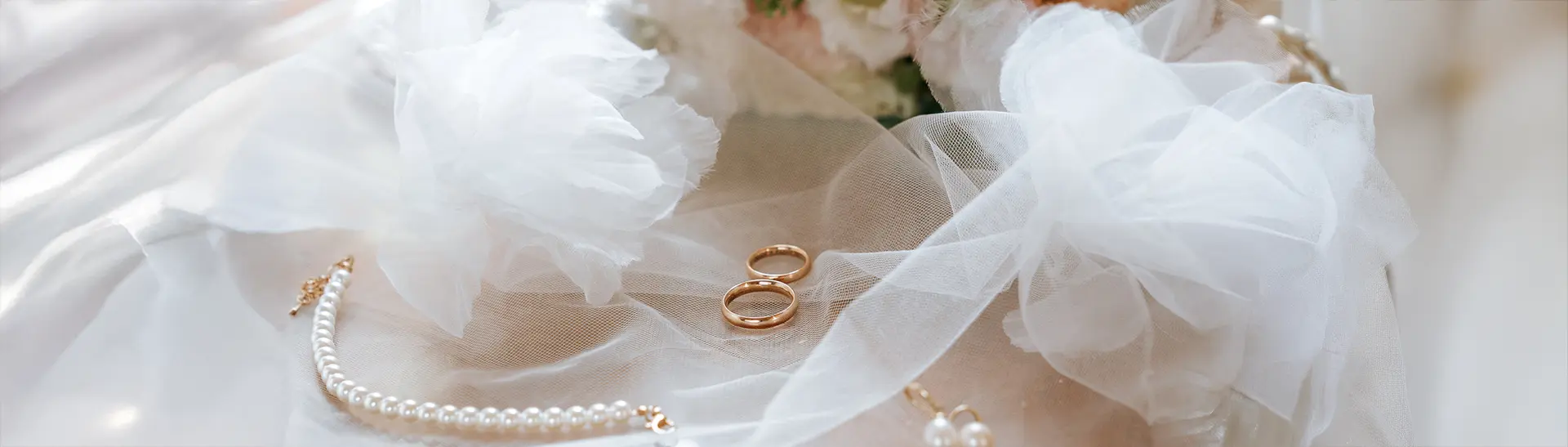 Weit arrangiertes Detailmotiv mit Ringen auf Tüll, daneben Perlenkette und sanftes Licht. Fine art hochzeitsfotografie trifft moderne hochzeitsfotos und schafft elegante Akzente