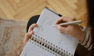 This is a GIF of a person seated and writing in a spiral notebook. The notebook is resting on their lap, and they are using a pen in their right hand to jot down notes or sketches. The view is from above, focusing on the notebook and the person's hands, with the patterned floor in the background. The motion is repetitive, showing the person writing, pausing to think, and then continuing to write.