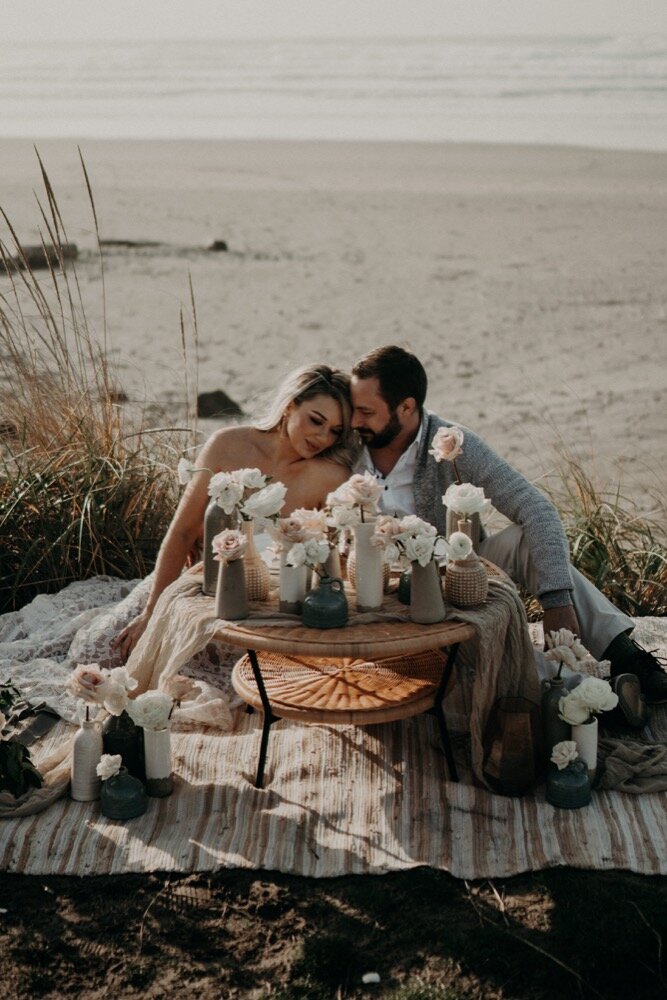 A couple sits on a beach picnic setup surrounded by vases of flowers, leaning affectionately into each other.
