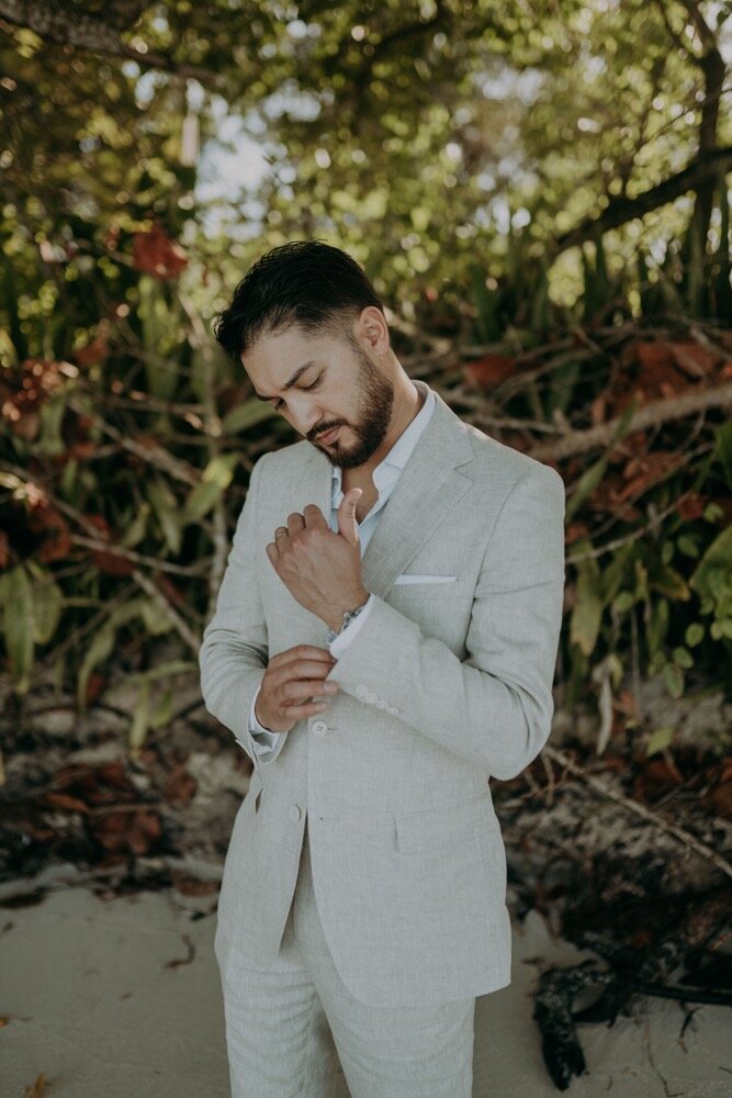 Groom in light suit adjusting his cufflinks outdoors with greenery behind him.