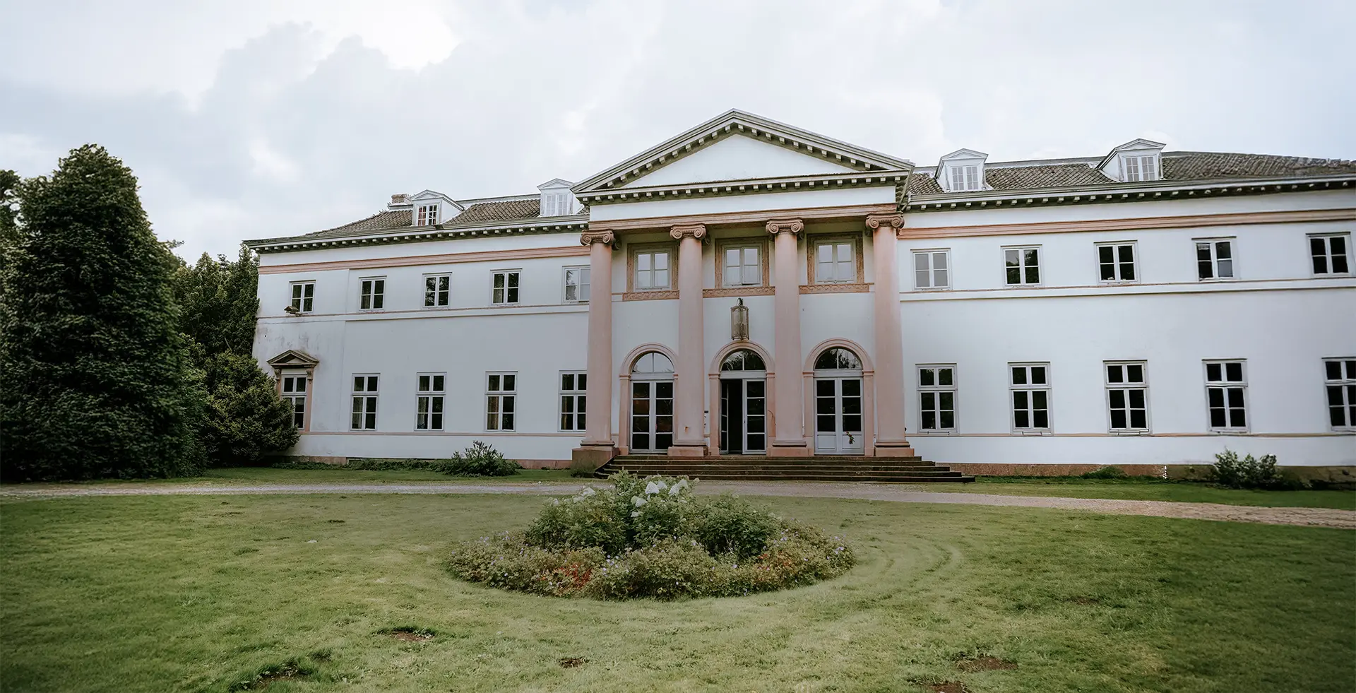 Weißes Herrenhaus mit rosafarbenen Säulen, symmetrischen Fenstern und Rasenrondell – ein einladender Ort für eine Schloss-Hochzeit, perfekt eingefangen in Hochzeitsfotografie mit Stil