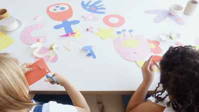 Two young girls sitting at a table cutting colorful paper and working on an art project