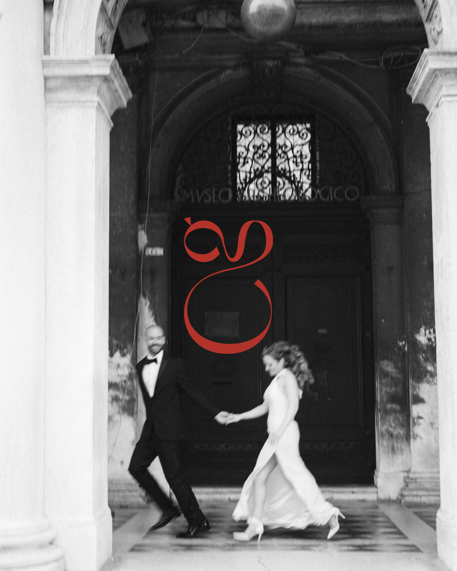Black and white photo of a couple joyfully walking hand-in-hand under an arched hallway, overlaid with a bold red brand mark.