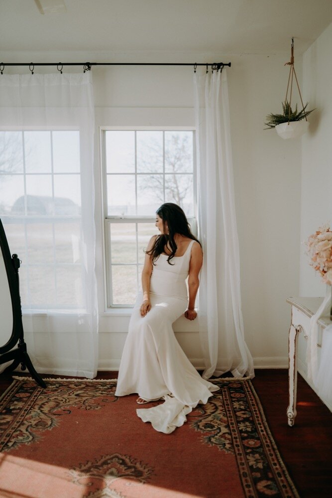 Bride sitting on a windowsill in a white gown, looking out at the landscape