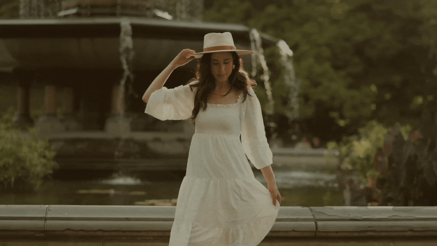 Founder Ariana of Ivy Grows Studio in a white summery dress and straw hat spinning in a circle and holding on to her hat next to the fountain
