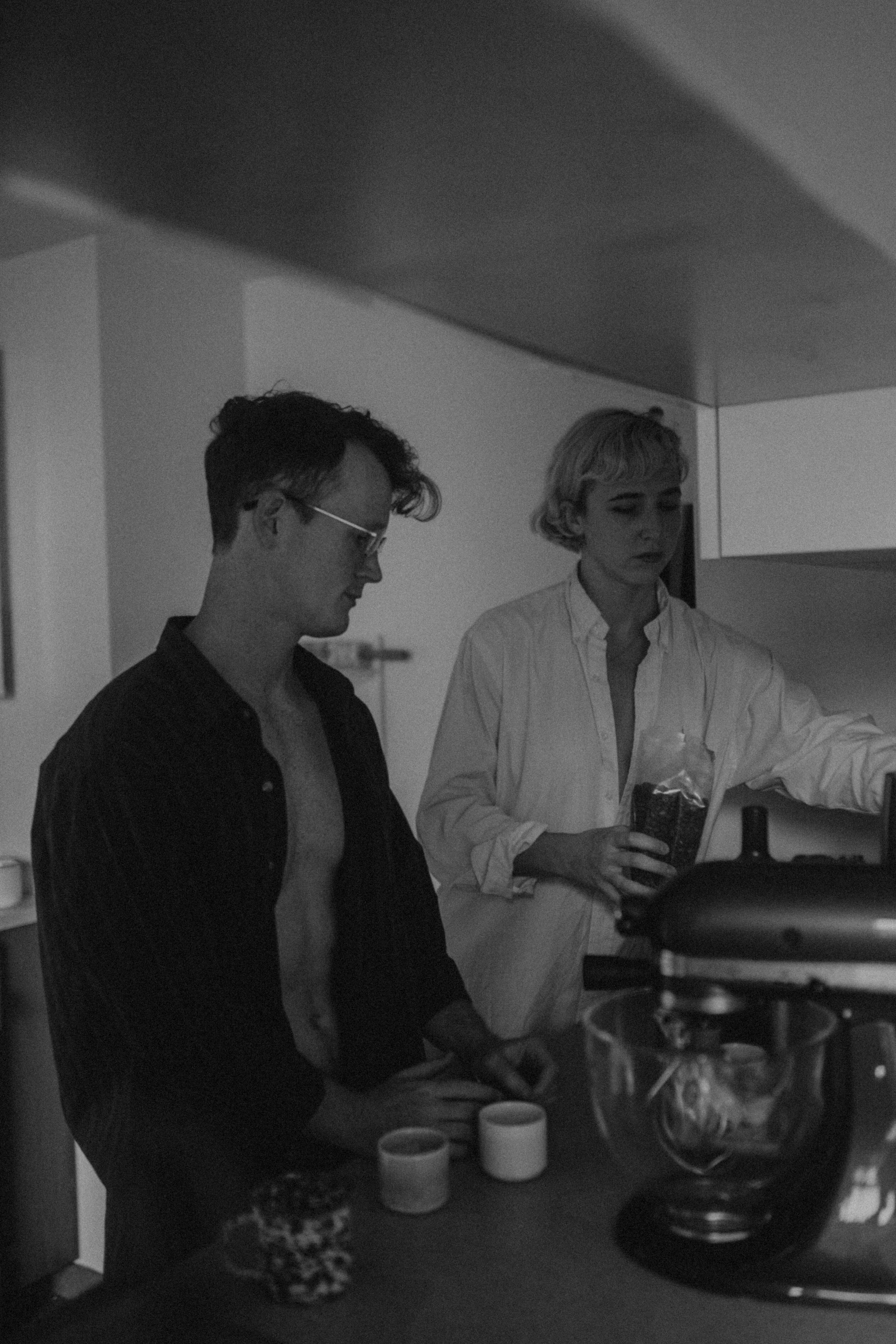 A couple, both wearing oversized shirts, stands in a kitchen making coffee in soft black and white tones.