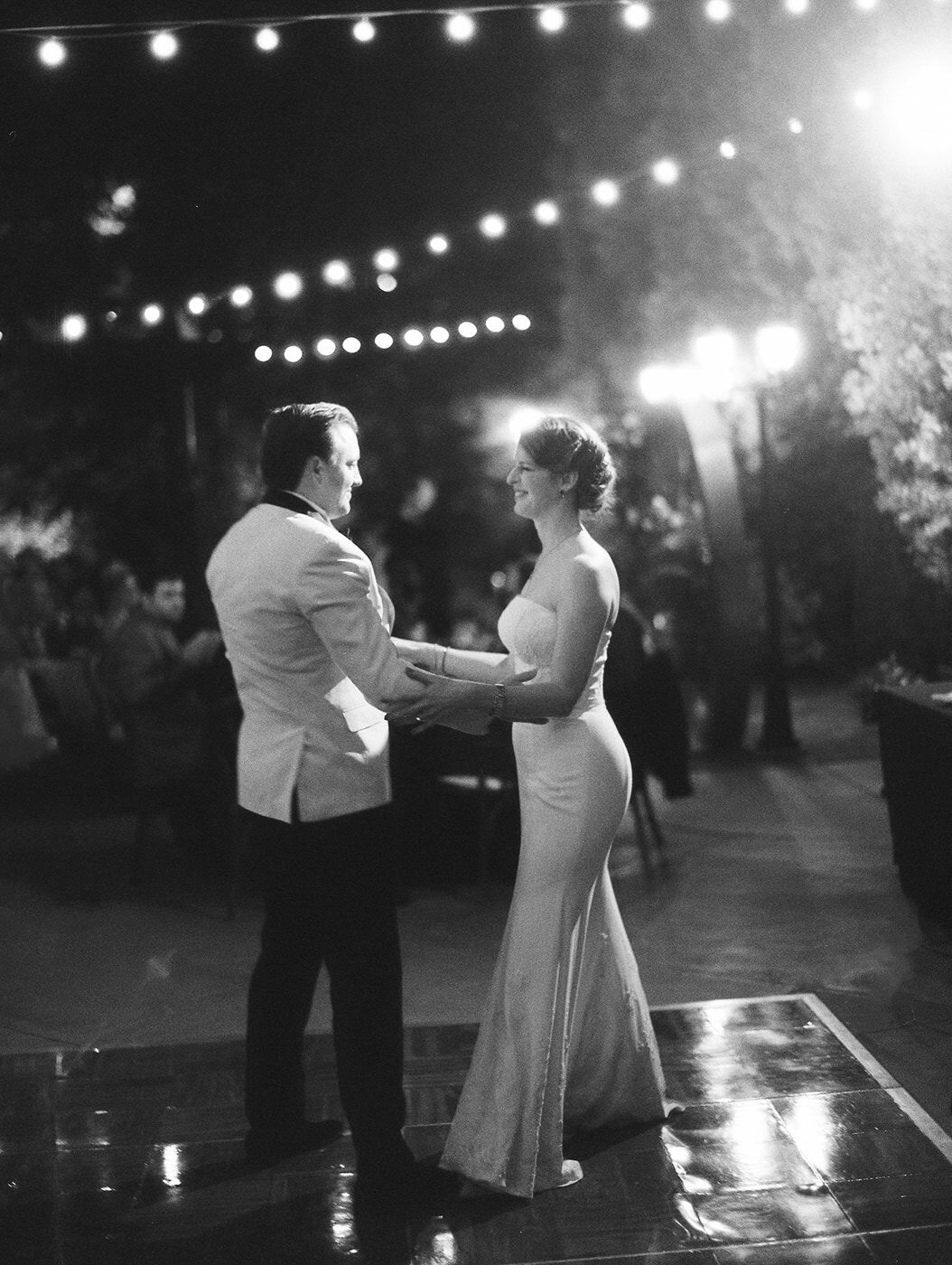 a bride and groom during their first dance at Franciscan Gardens in San Juan Capistrano. There are market lights above and behind them, they are smiling at eachother, and the image is on black and white film and is a little grainy.
