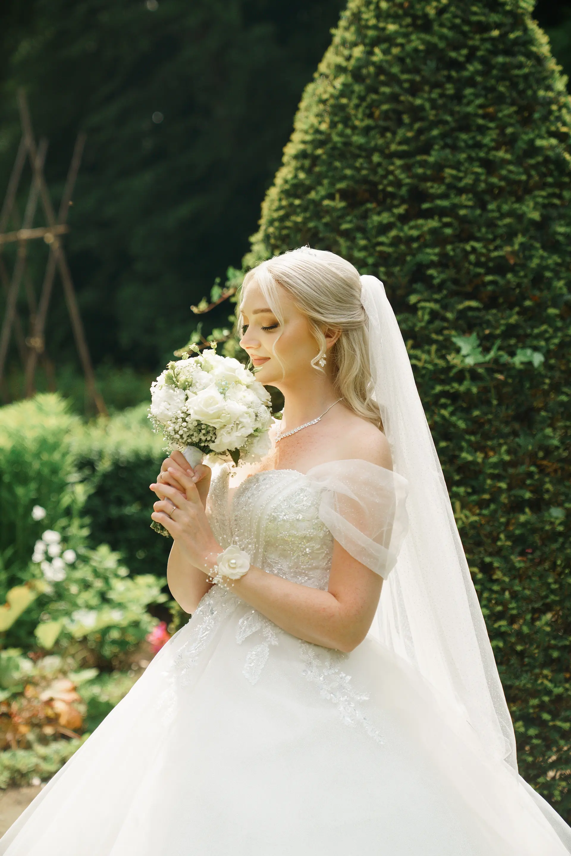 Braut mit weißem Strauß vor geschnittener Hecke; weiches Gartenlicht betont natürliche hochzeitsfotografie, emotionale hochzeitsbilder und hochzeitsfotografie mit stil
