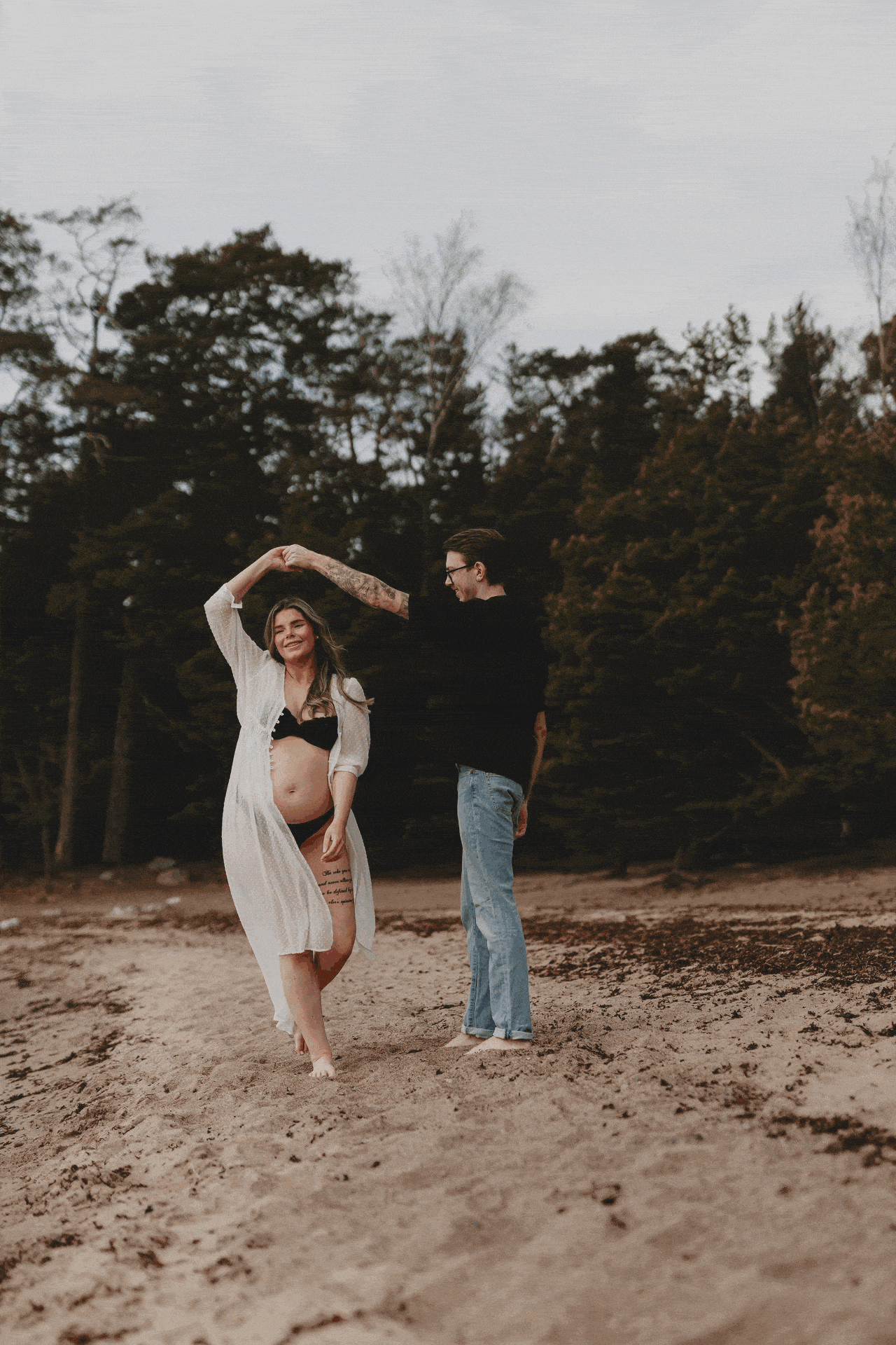 En gravid kvinna tillsammans med sin partner dansandes på en strand under en gravidfotografering i Stockholm
