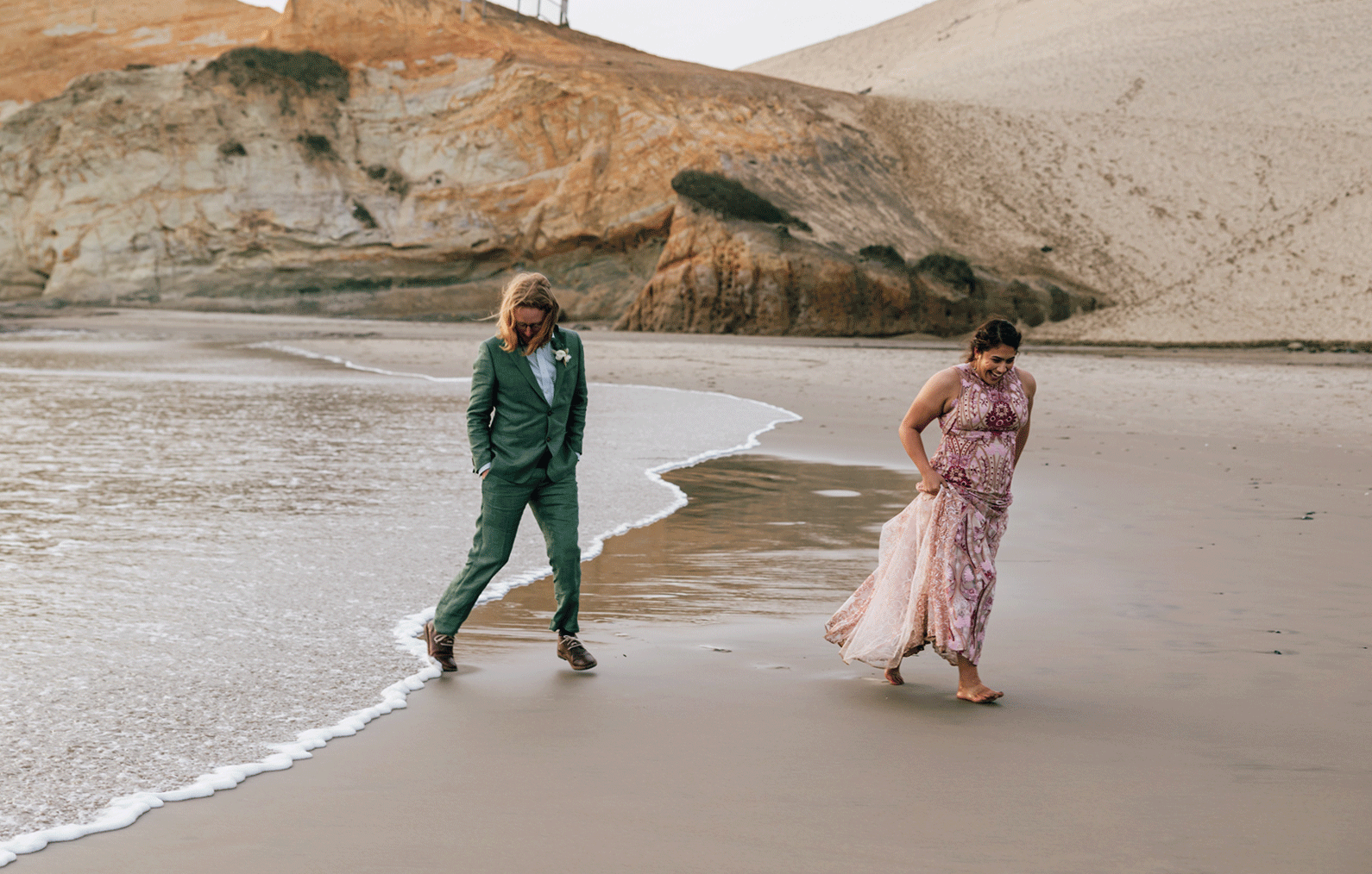 A bride in a pink Rue de Seine gown holds up her dress while running barefoot on the beach while her groom in a green suit runs from the ocean waves. 