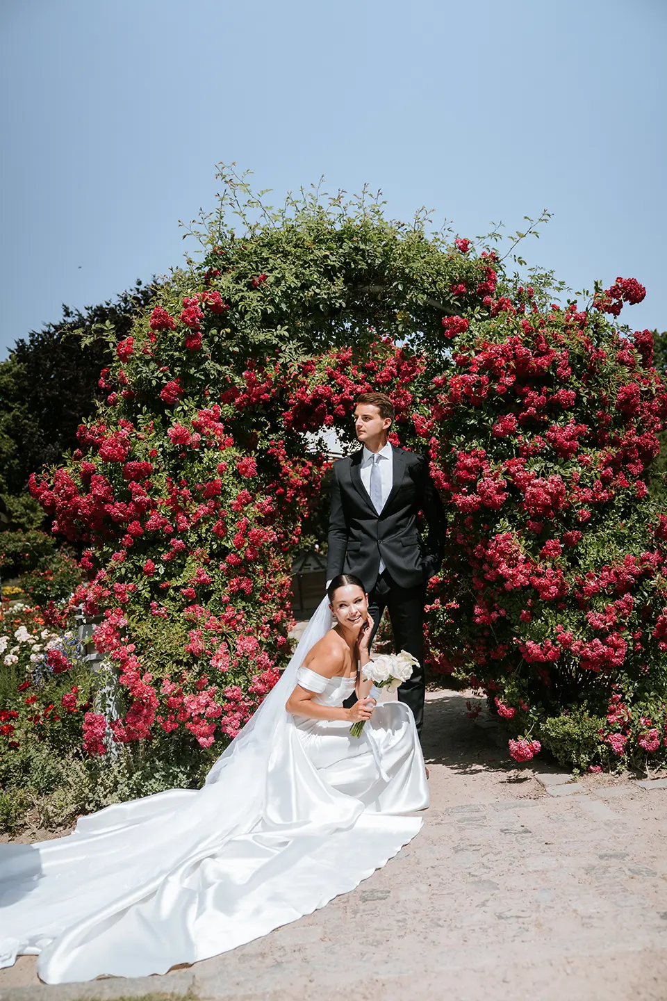 Vor einem großen Rosenbogen kniet die Braut mit langer Schleppe und Bouquet, der Bräutigam steht dahinter; hochzeitsfotografie mit stil und emotionale hochzeitsbilder