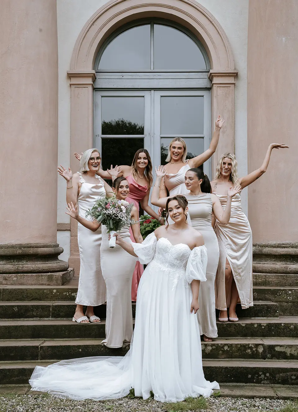 Braut in elegantem Kleid mit sechs Brautjungfern auf der Treppe vor historischer Fassade; vertikales Mobil-Header-Bild – natürliche Hochzeitsfotografie, hochzeitsbilder draußen