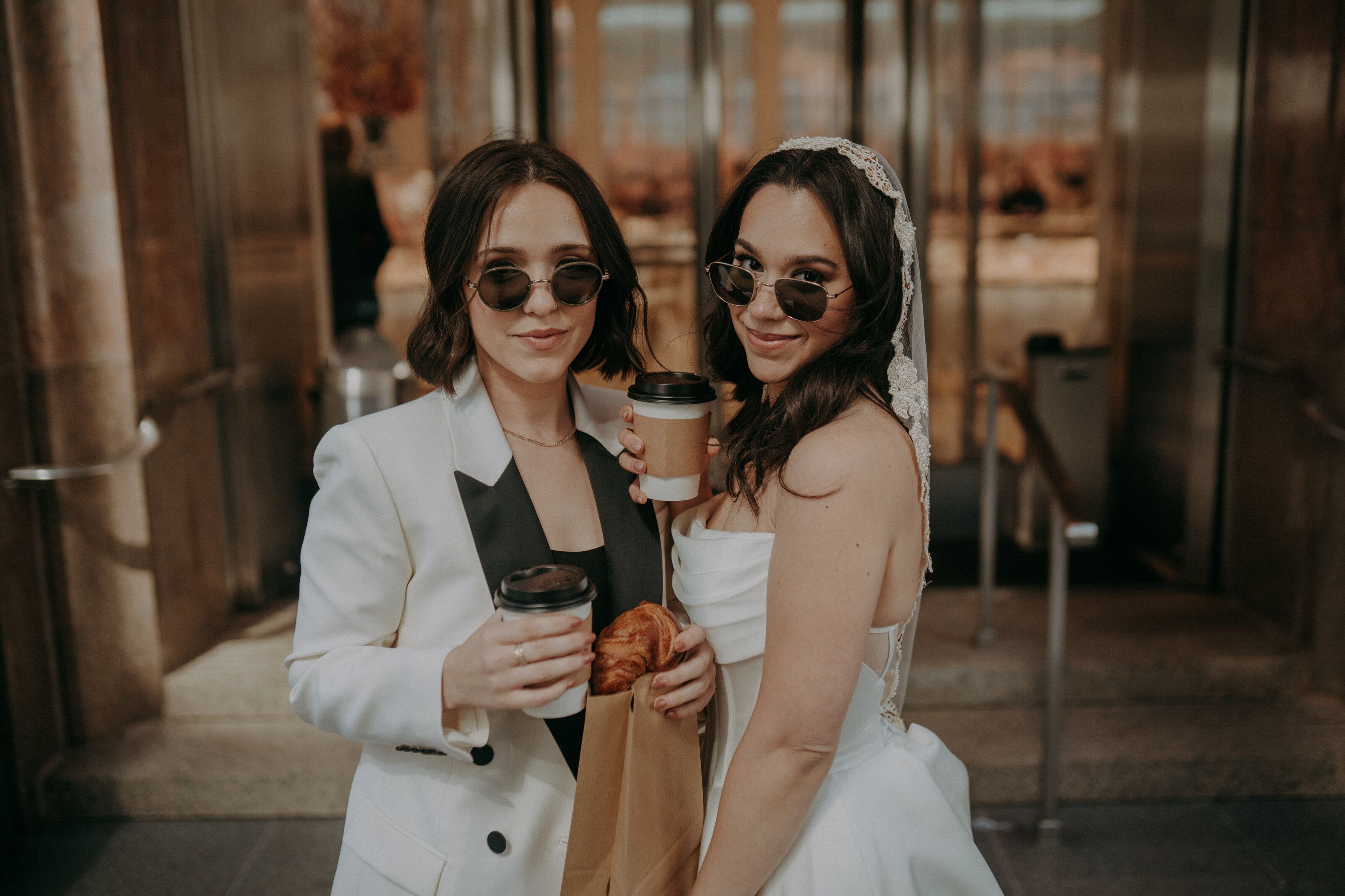 Two brides standing outside Tiffany & Co. wearing wedding attire and sunglasses, holding coffee and pastries with joyful expressions.