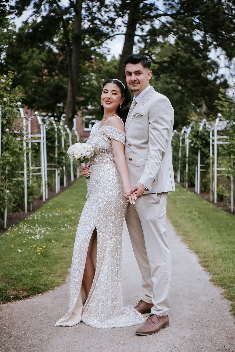 Das Paar posiert Hand in Hand auf einer von Spalieren gesäumten Allee, die Braut trägt einen weißen Strauß; moderne hochzeitsfotografie und fotograf hochzeit premium