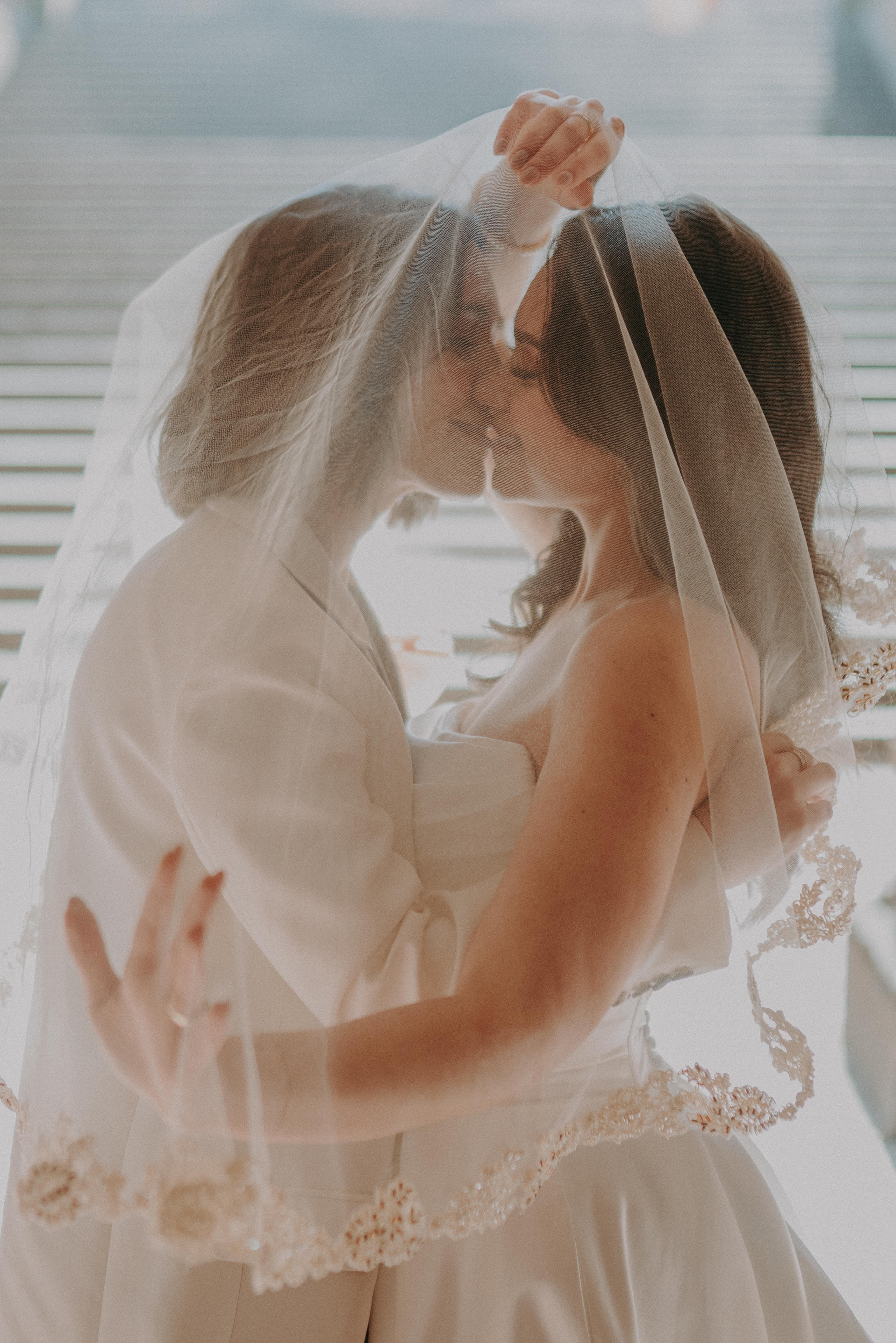 Two brides share a kiss under a wedding veil, faces close and softly lit.