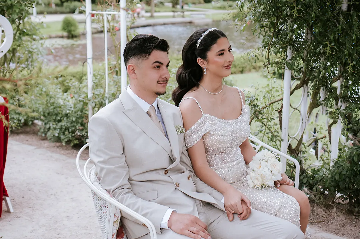 Braut und Bräutigam sitzen Hand in Hand unter Rosenbögen am Teich; beiger Anzug, funkelndes Kleid – natürliche hochzeitsfotografie und emotionale hochzeitsbilder