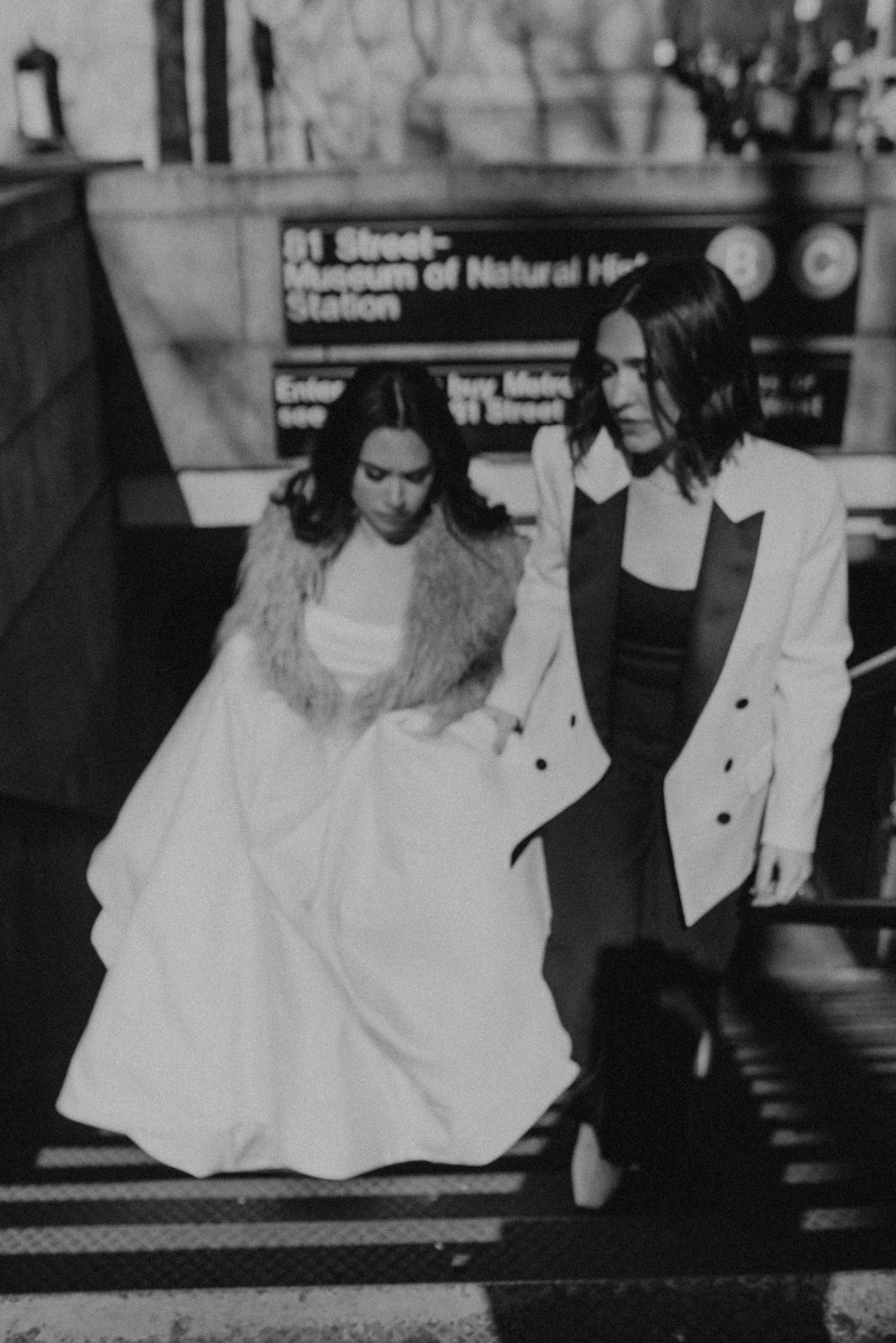 A bride in a white gown is helped up the stairs of a subway station by her partner in a white blazer, both in black and white.