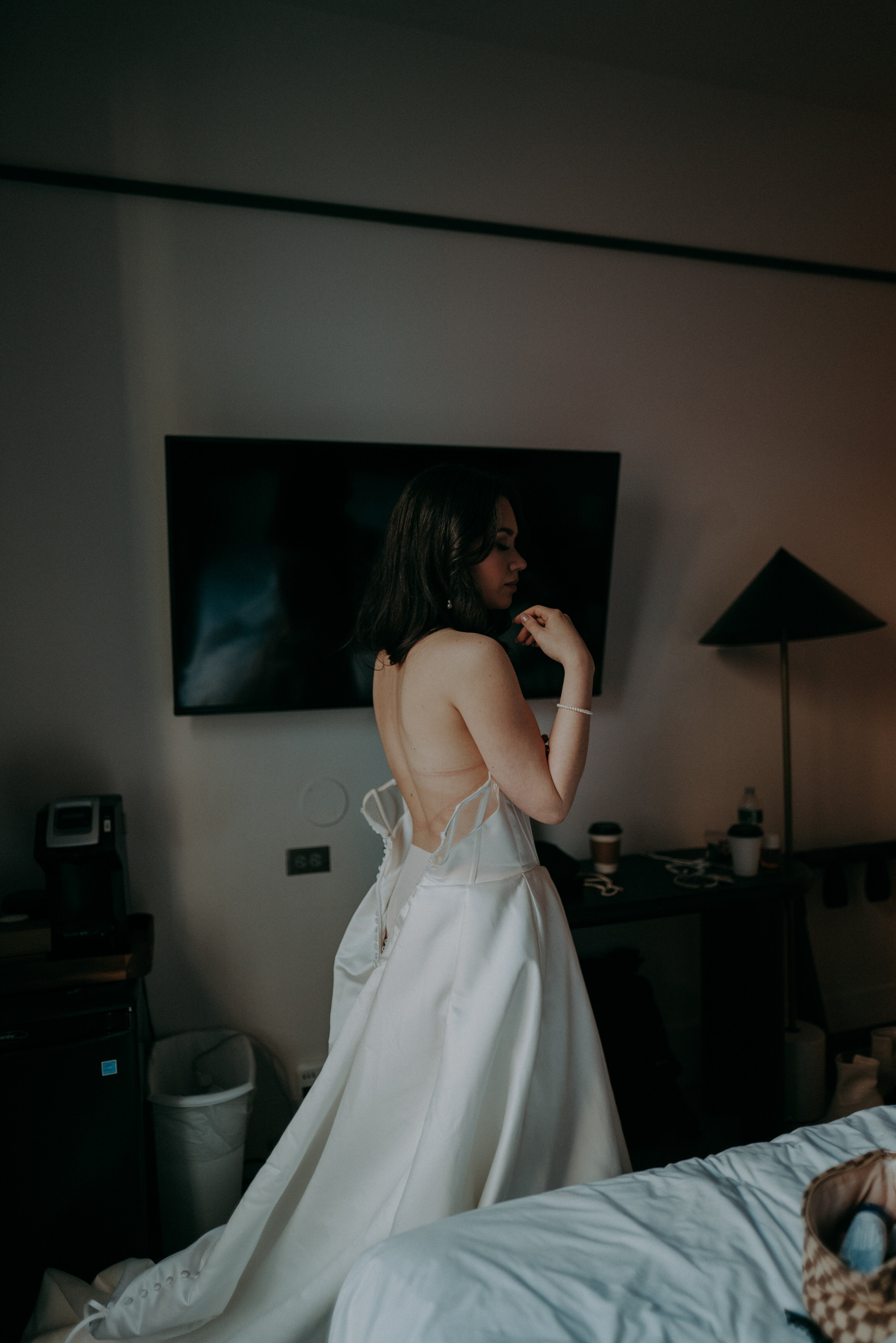 Bride in a backless wedding dress adjusting her hair in a dimly lit hotel room, with a bed and television in the background.