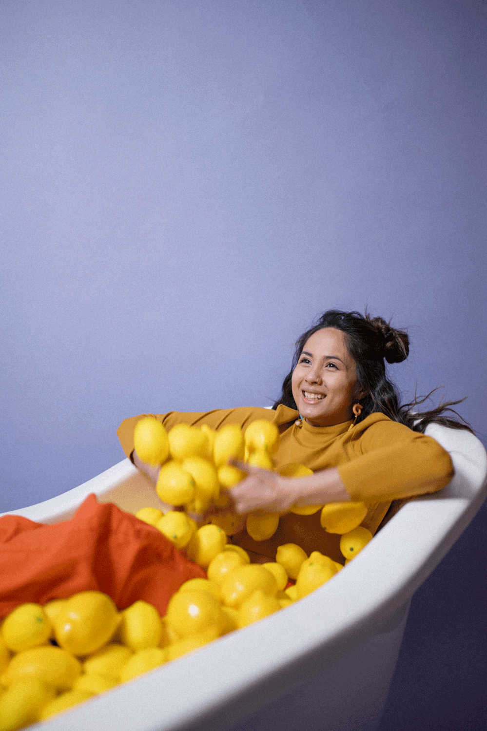 GIF of young Filipino woman wearing a mustard top and orange pants sits in a bathtub throwing lemons in the air and smiling in front of a purple backdrop.