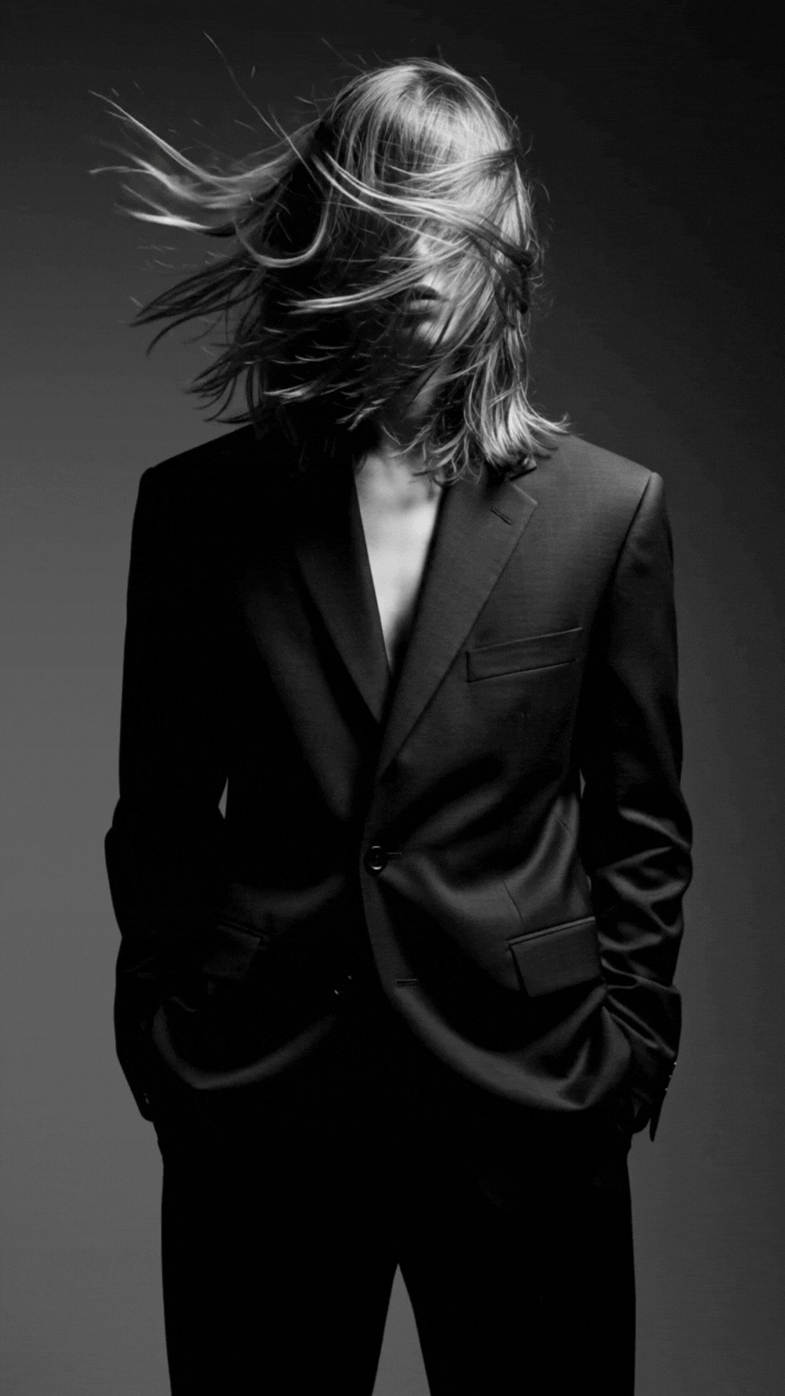 A female standing in a black suit with her hair blowing in over her face.