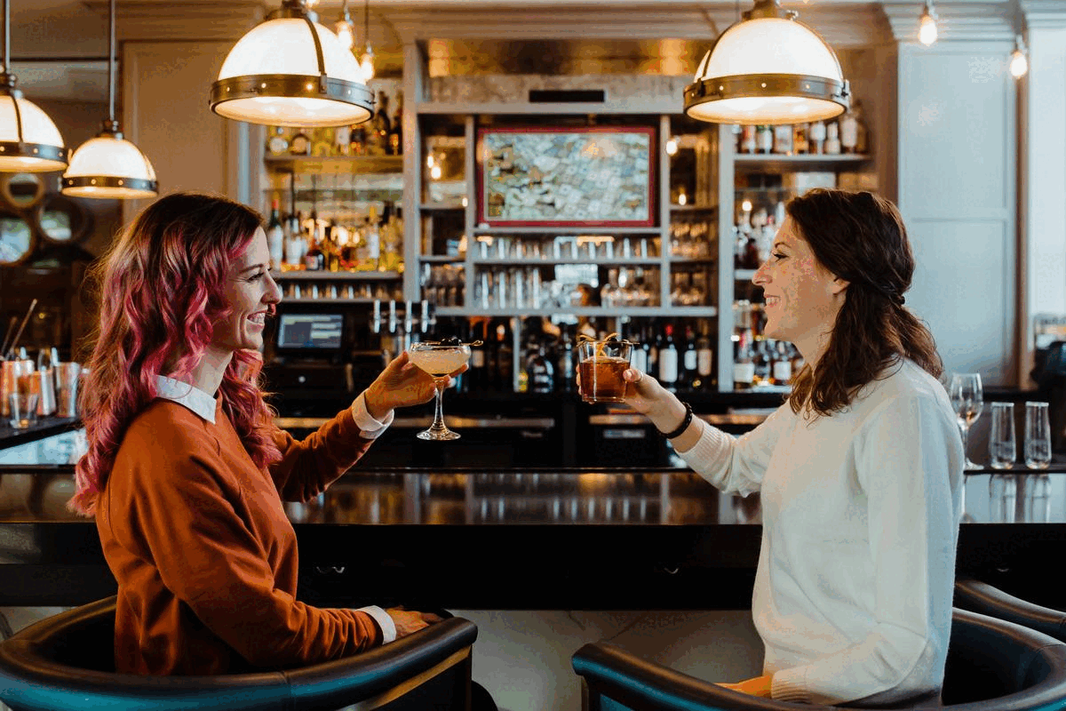 two women clinking glasses while sitting at bar