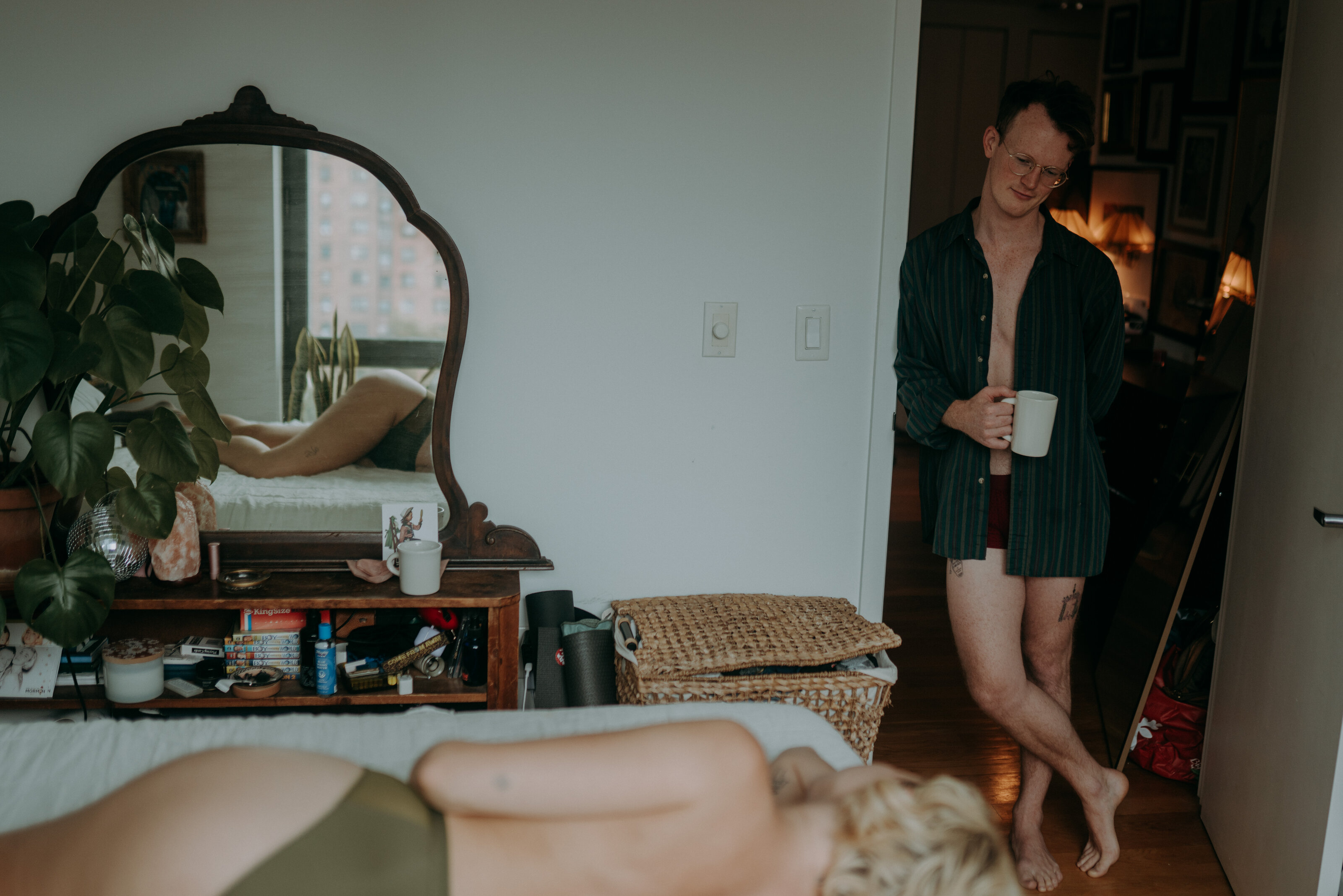 Man leaning in doorway with mug, looking at partner lying on bed, intimate morning scene.
