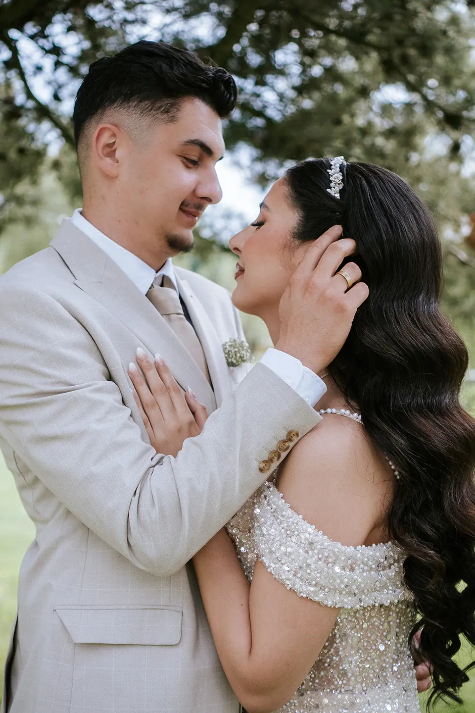 Der Bräutigam hält die Braut sanft am Gesicht, beide lächeln und zeigen ihre Ringe unter dichtem Blätterdach; emotionale hochzeitsbilder und hochzeitsfotografie mit stil