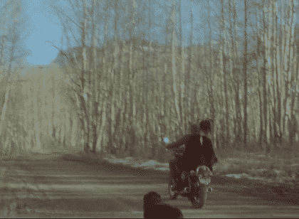 A video of a couple on a motorcycle driving down a dirt road in the winter. A black dog is following them.