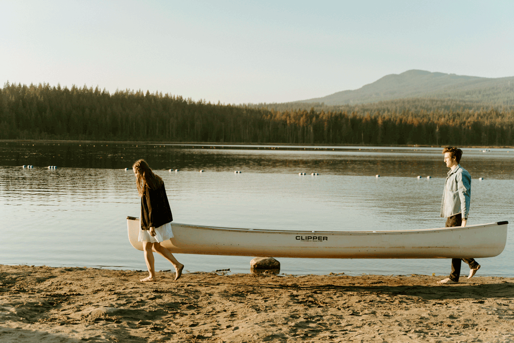 gif of a girl and boy carrying a canoe across the beach at a lake