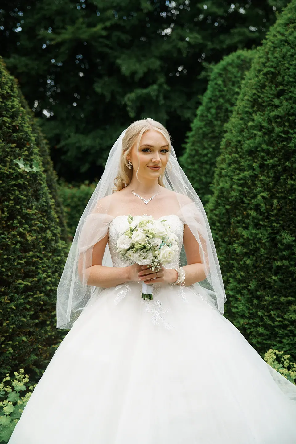 Braut in schulterfreiem Kleid hält einen weißen Rosenstrauß zwischen geschnittenen Hecken; sanftes Licht und ruhiger Ausdruck – natürliche hochzeitsfotografie und fine art hochzeitsfotografie