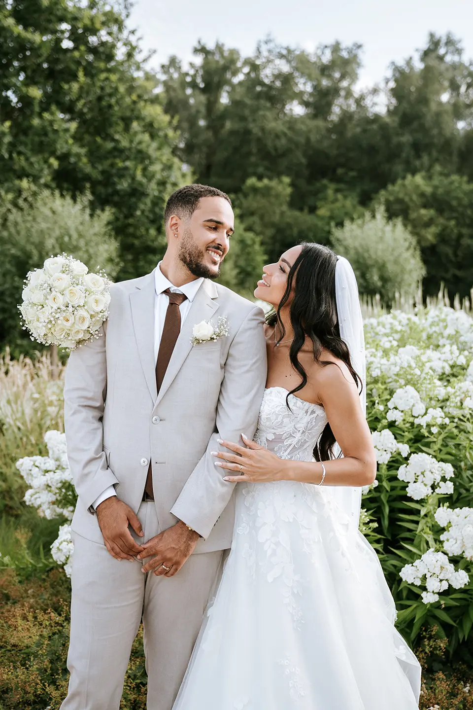 Das Paar lächelt sich vor weißen Blüten an, der Strauß ruht lässig auf seiner Schulter. Moderne Hochzeitsfotografie mit natürlichen Hochzeitsbildern in sanftem Abendlicht