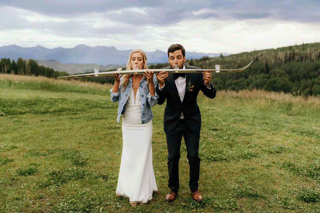 bride and groom doing shots off a ski with mountains in the background