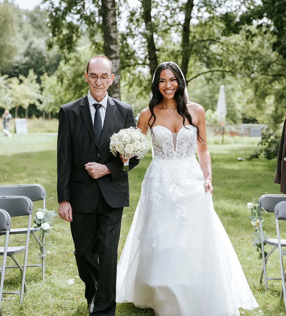 Zwischen Reihen von Gästen schreitet die Braut am Arm ihres Vaters über die Wiese, weißer Rosenstrauß in der Hand; reportagefotografie hochzeit, ganztägige hochzeitsreportage und natürliche hochzeitsfotografie