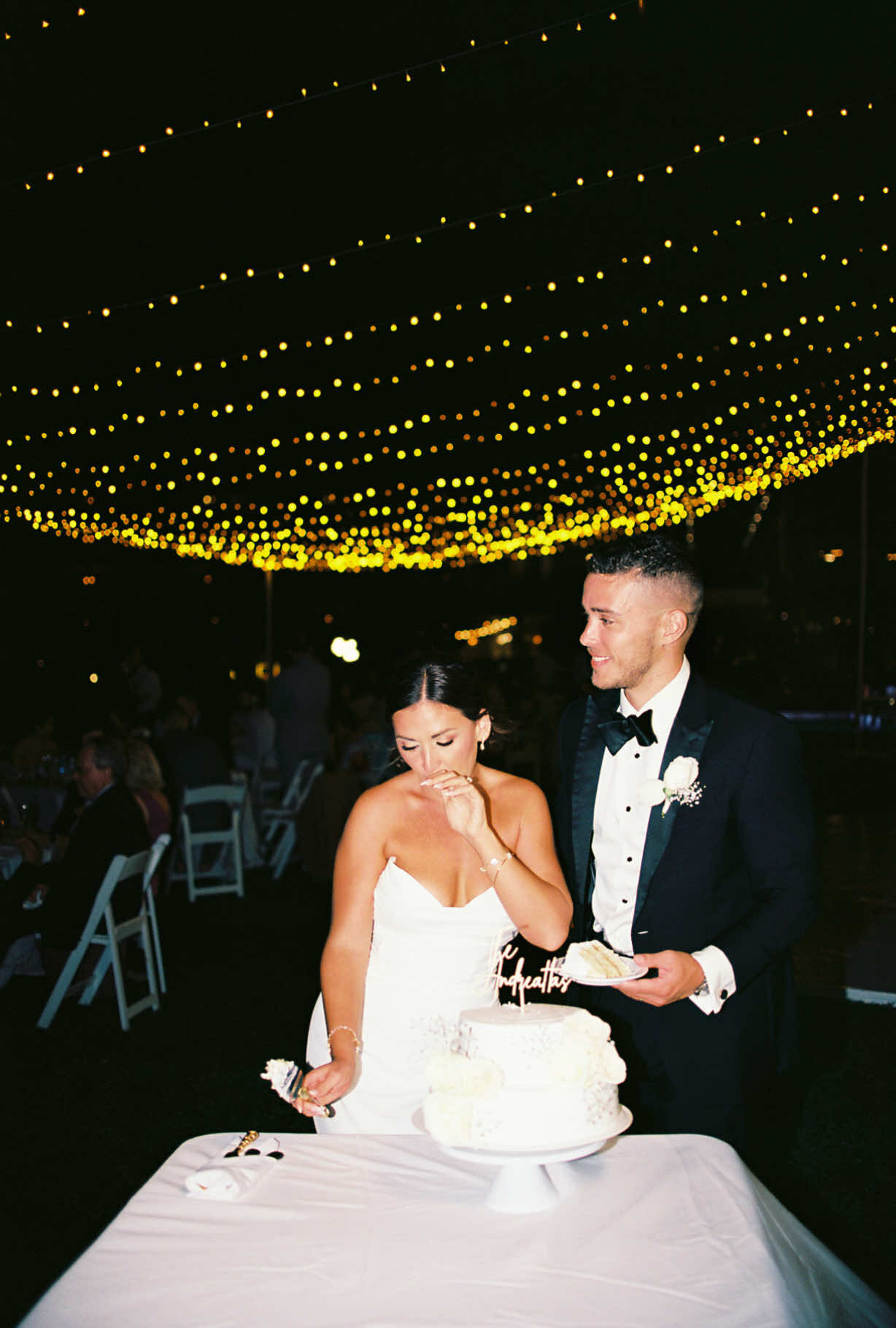 A man and a woman are standing next to each other, likely at a wedding reception. The woman is wearing a bridal gown, while the man is dressed in formal attire or a suit