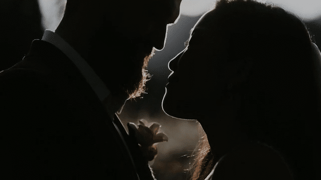 A video of a bride and groom standing in a meadow with mountains in the background. The video starts with a far off arial shot of them and moves closer until it cuts to a closeup of their faces.