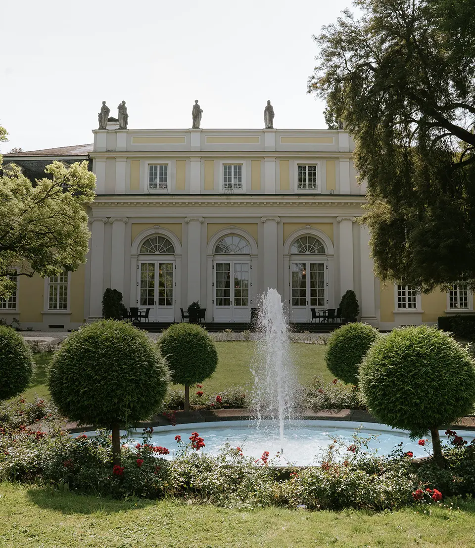 Vor der klassizistischen Villa sprudelt ein Brunnen, umrahmt von Rosen und Buchskugeln – perfekte Kulisse für natürliche Hochzeitsfotografie und hochzeitsbilder draußen