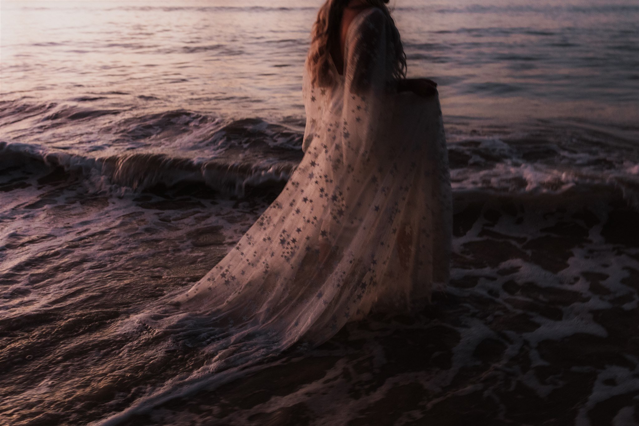 a bride wearing a beautiful sheer wedding gown with reflective stars shimmering in a purple sunset as she wades through waves