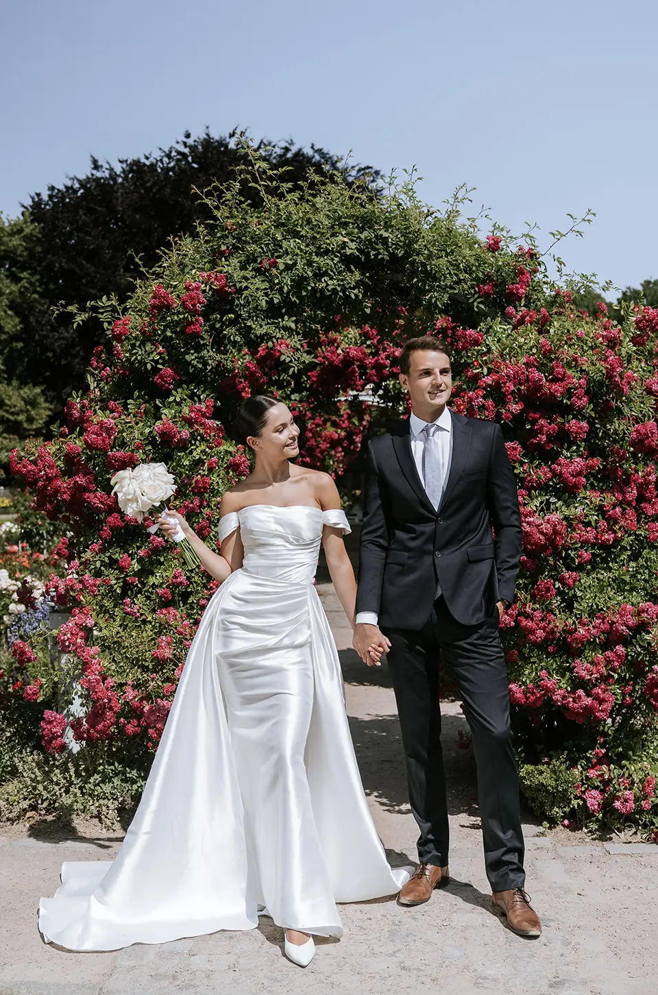 Braut im Satin-Off-Shoulder-Kleid und Bräutigam im Anzug Hand in Hand vor üppigem Rosenbogen; natürliche Hochzeitsfotografie mit stilvollen Hochzeitsbildern draußen