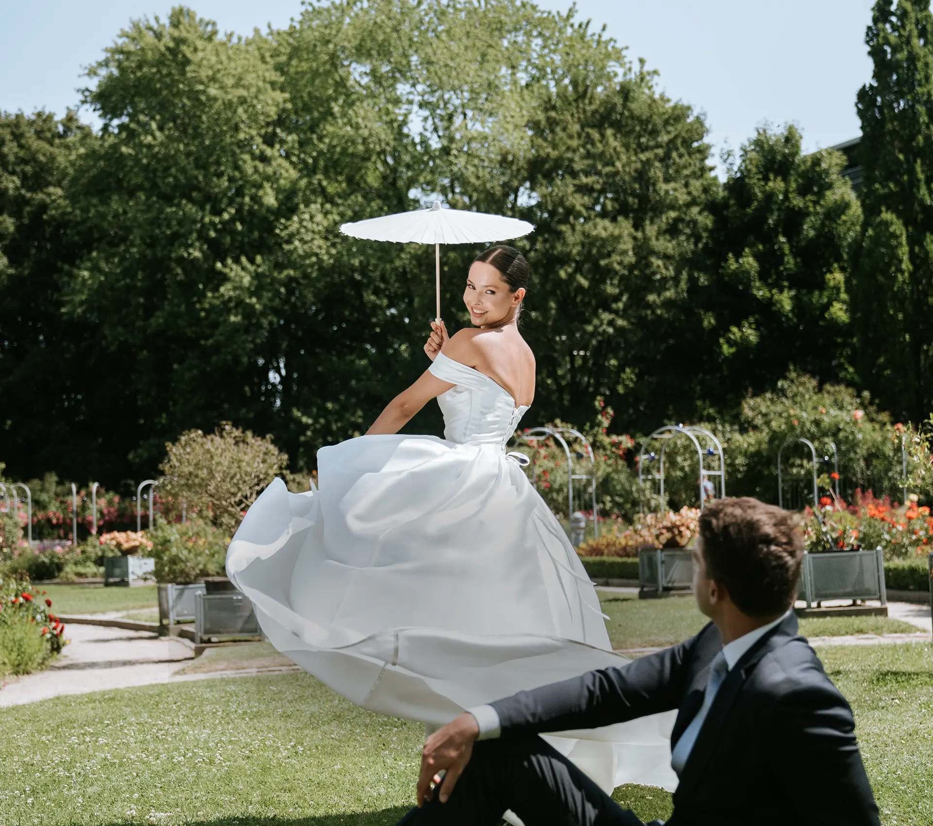 Braut dreht sich mit weißem Schirm, Bräutigam sitzt im Park; lebendige Sommerhochzeit in Münster – natürliche Hochzeitsfotografie, hochzeitsbilder draußen