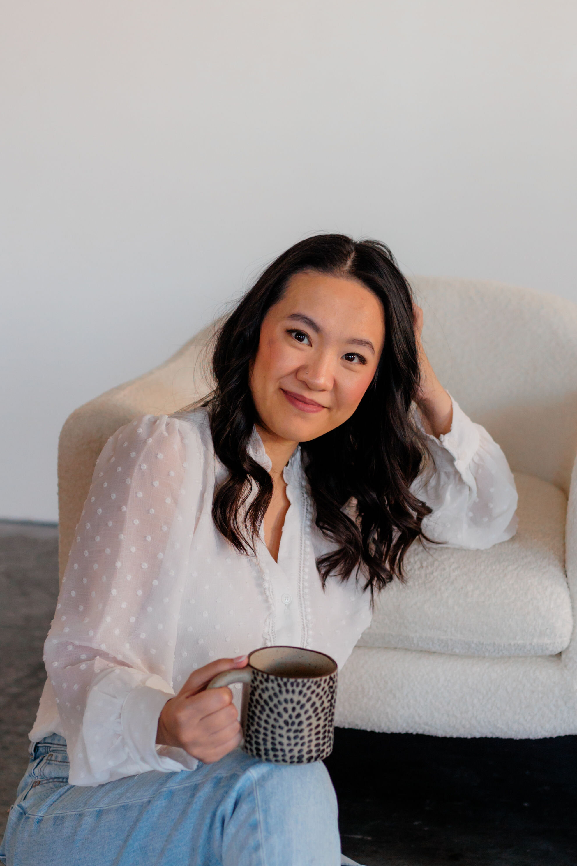 Woman with long dark hair wearing a white blouse sits on the floor next to a cream-colored chair, holding a mug and smiling at the camera—perfectly capturing the warm, approachable vibe of expert branding and website design.