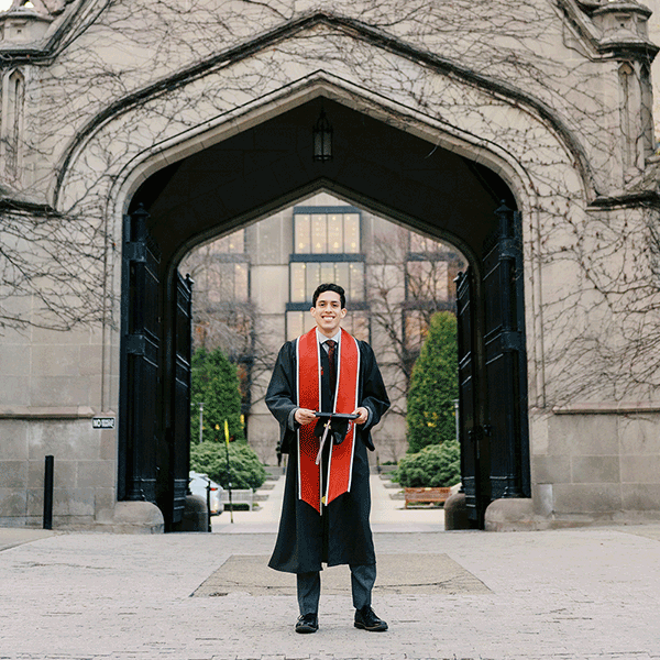 Senior guy wearing cap and gown and leaning against a brick wall
