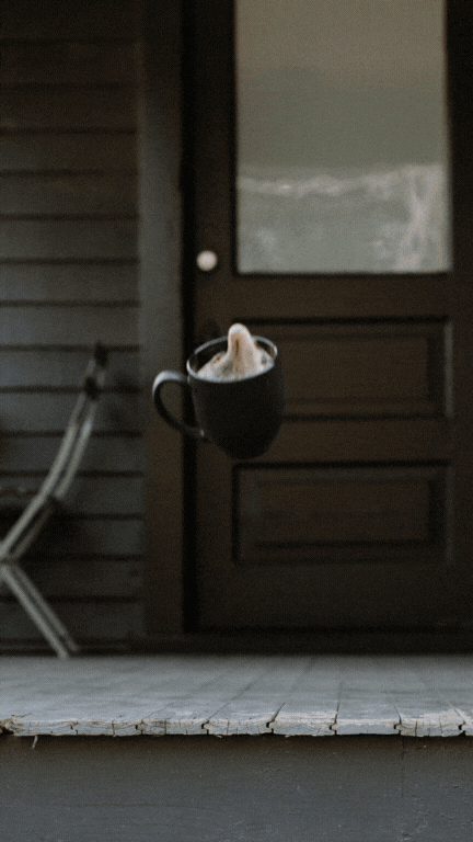 A black mug floats in midair, hot steam rising from it, in front of a dark wooden door—perfect inspiration for anyone looking to become a white label designer. A porch with a folded chair is visible on the left.