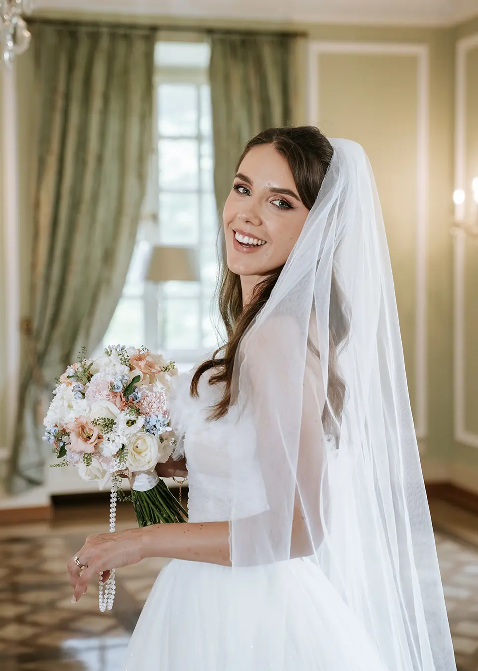 Lachende Braut im Salon, der Schleier rahmt das Gesicht, pastellfarbener Blumenstrauß in der Hand; moderne hochzeitsfotografie und hochzeitsfotografie mit stil
