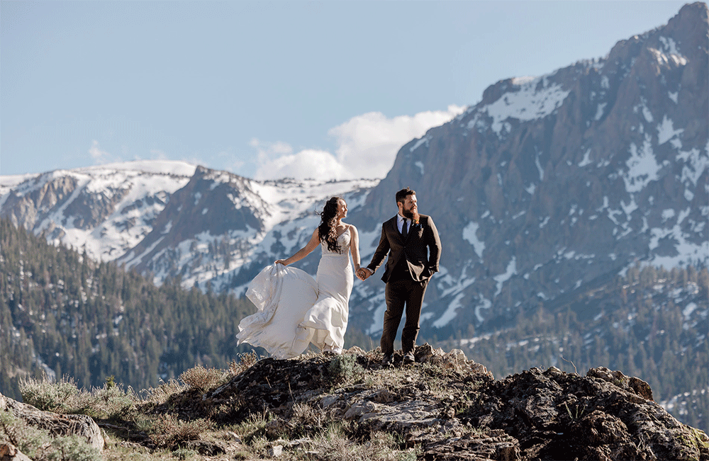 A bride and groom stand in front of snow capped mountains on a their very windy mammoth wedding day. 